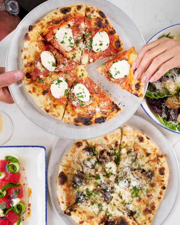 A table of pizzas and salads with lemon drinks; hands share a slice while toppings include mozzarella and herbs, delicious vibes.