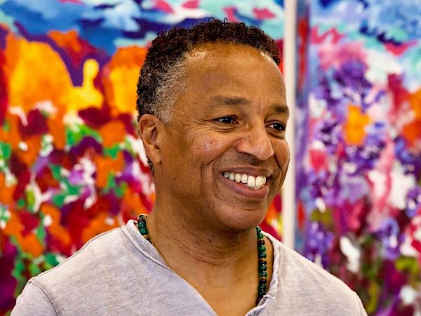 A person smiling, standing in front of colorful abstract paintings. The scene is vibrant with red, blue, and purple hues in the background.