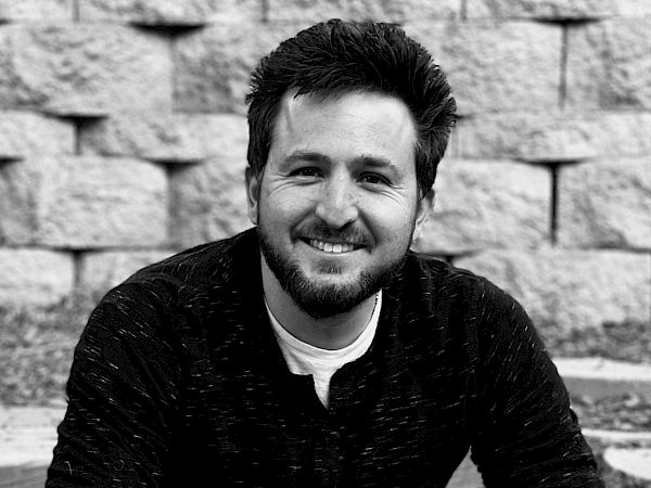 A person sitting and smiling, with a stone wall in the background. The image is black and white.