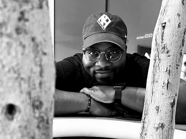 A person wearing a cap and glasses is resting their arms on a surface, with rocks and tree trunks in the foreground.
