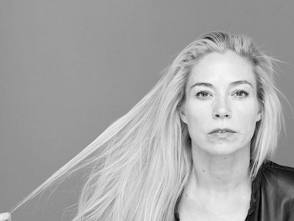 A person in a black outfit poses in a monochrome photo, holding their long, straight hair with a focused expression, set against a plain background.