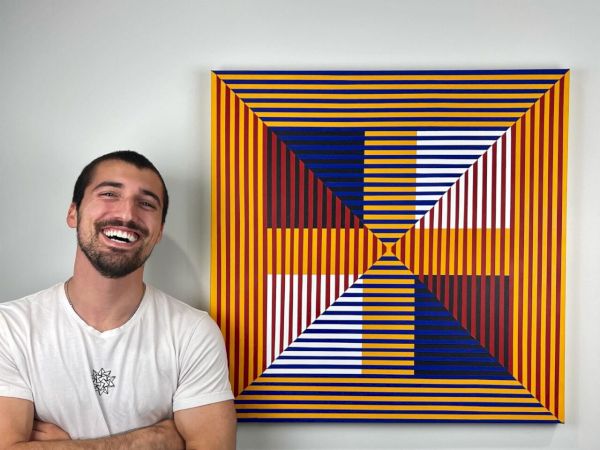 A person stands smiling with arms crossed beside a colorful geometric artwork featuring stripes and squares on the wall.