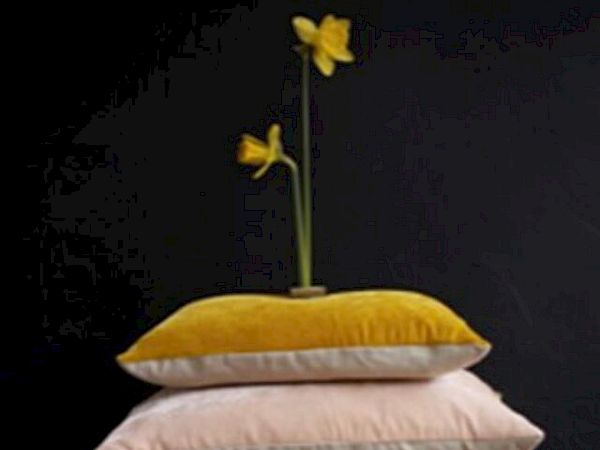 A stack of colorful pillows in shades of yellow, pink, and orange with two yellow flowers placed on top against a dark background.