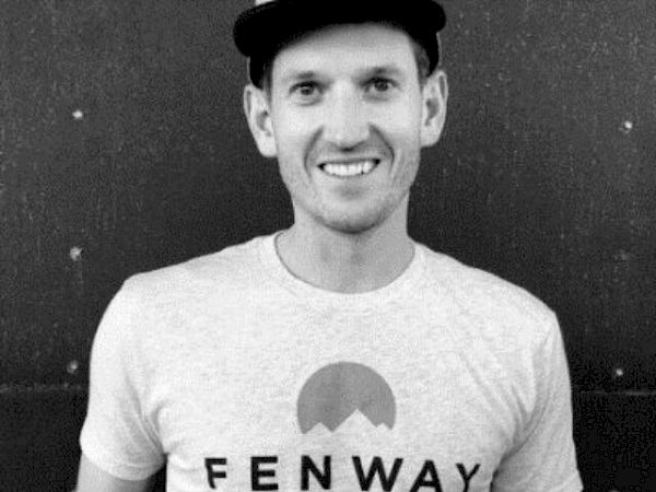 A person wearing a "Fenway Clayworks" T-shirt and cap, holding an object, smiles at the camera in a black-and-white photo.