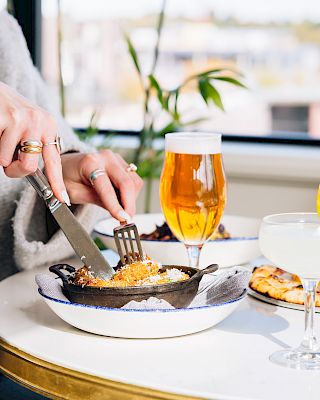 A person is cutting into a pasta dish at a bright restaurant table with a beer, a glass of water, and lemon wedges nearby, enjoying a meal.