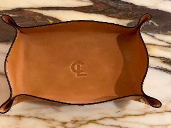 A brown leather tray with curled edges is placed on a marble surface. The tray features embossed initials in the center.