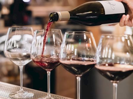 A person is pouring red wine into a row of five wine glasses on a countertop in a well-lit setting.