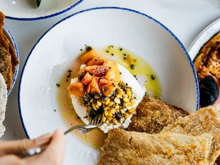 A dish with soft cheese, diced tomatoes, seeds, seasoned oil, and flatbread. Nearby, there's a salad with veggies and another dish visible.