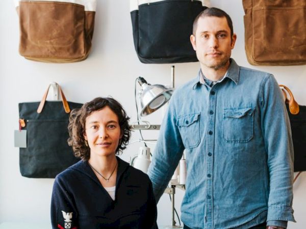 A man and woman stand in a room with tote bags displayed on the wall behind them.