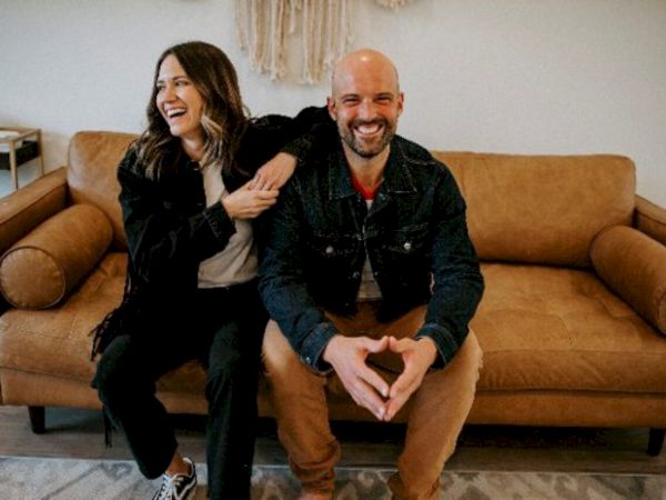 A man and woman are sitting on a brown couch, smiling and appearing relaxed in a cozy setting.