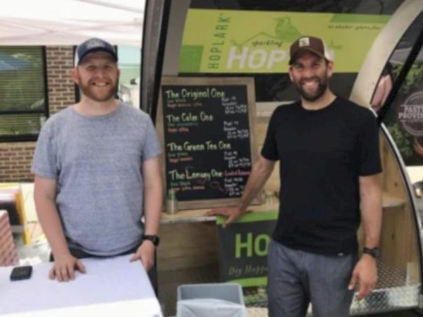 Two people stand at a kiosk with a menu board listing different items. They appear to be at a casual outdoor setting.
