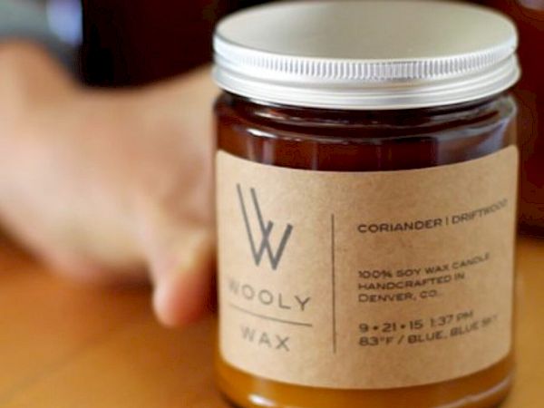 A glass jar labeled "Wooly Wax," featuring coriander and eucalyptus scent, with 100% soy wax, handcrafted in Denver, CO.
