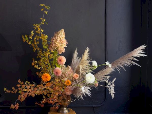 An elegant floral arrangement featuring pampas grass, orange and white flowers, and green foliage against a dark background.