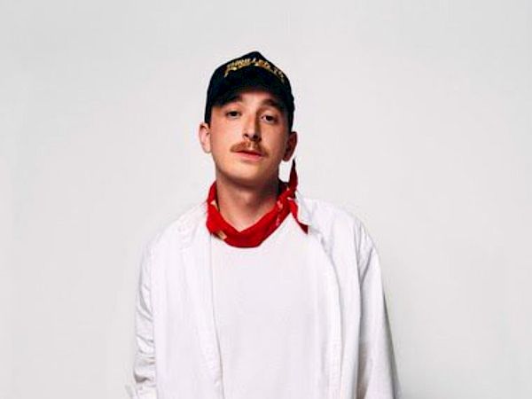 A person wearing a white shirt, red bandana, and cap, posing against a plain background.