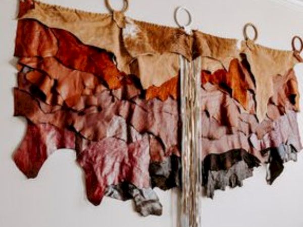 The image shows a wall-hanging art piece made of layered, textured fabric in earthy tones, arranged in a symmetrical pattern with circular accents.