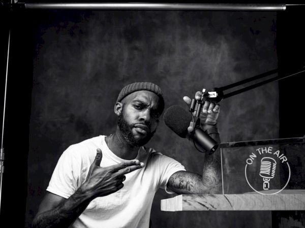A person in a studio setup pointing at a tattoo, with a microphone and "On the Air" sign beside them.