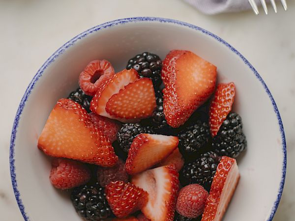A bowl of fresh mixed berries (strawberries, raspberries, and blackberries) on a light table, with a fork and napkin nearby, ending here.