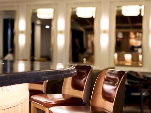 A chic bar area with tall wooden stools at a curved marble counter, warm pendant lights, and a softly lit lounge in the background, elegant and cozy.