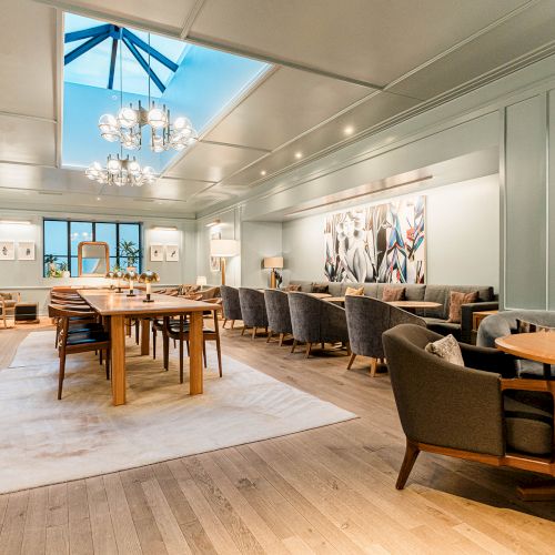 A bright, stylish dining or lounge area with wooden floors, round and rectangular tables, upholstered chairs, soft lighting, and a skylight chandelier.
