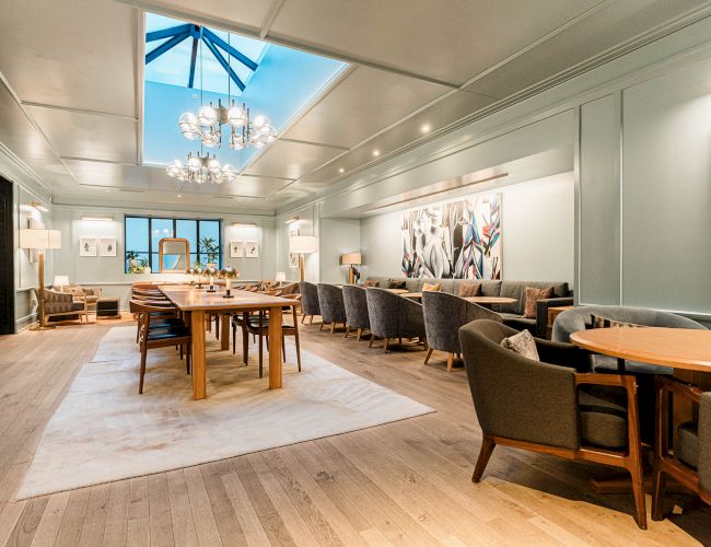 A bright, stylish dining or lounge area with wooden floors, round and rectangular tables, upholstered chairs, soft lighting, and a skylight chandelier.
