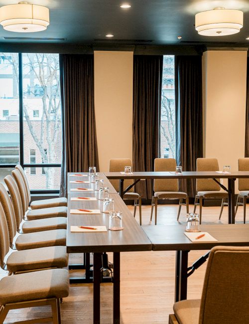 A conference room with long U-shaped tables, beige chairs, large windows, and soft ceiling lights, ready for a meeting.