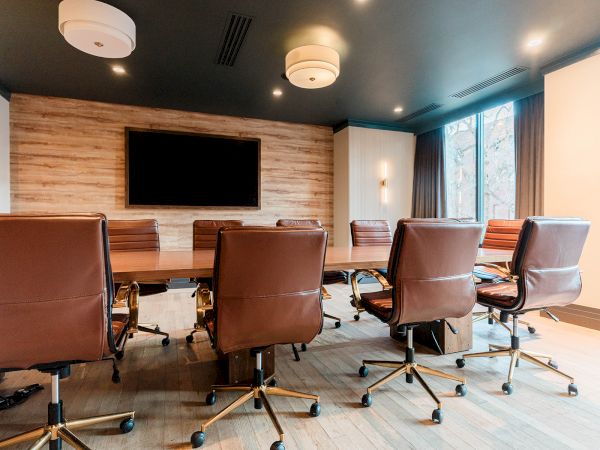 A modern conference room with a long wooden table, brown leather chairs, a wall-mounted TV, wood panel accent wall, and large windows letting in natural light.
