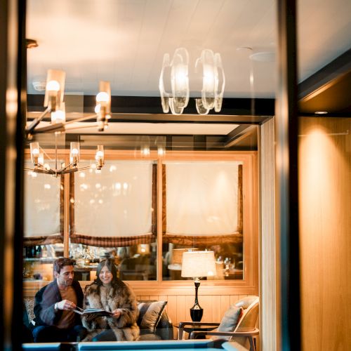 A cozy lounge viewed through a glass window: a couple sits on a couch with lamps, chandeliers, and warm lighting.