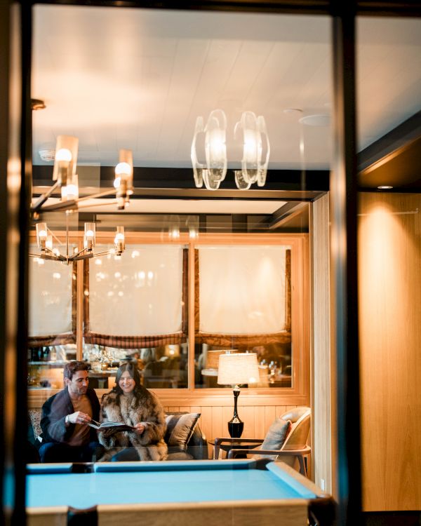 A cozy lounge viewed through a glass window: a couple sits on a couch with lamps, chandeliers, and warm lighting.