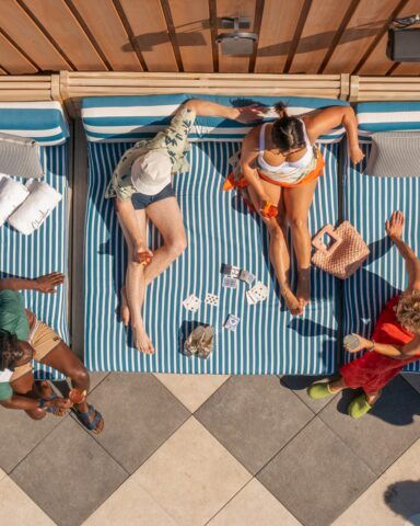Aerial view of four people relaxing on striped sunbeds with towels and drinks, in a checkered patio area, next to a wooden fence.