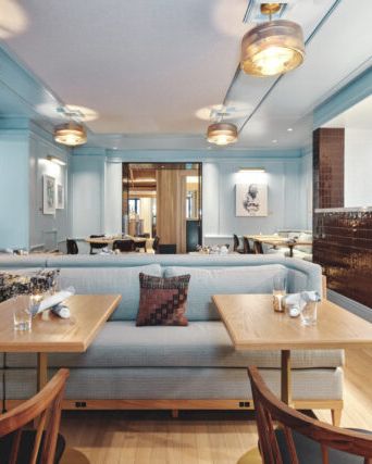 The image shows a modern restaurant interior with wooden tables, cushioned seating, stylish lighting, and a tiled bar area.