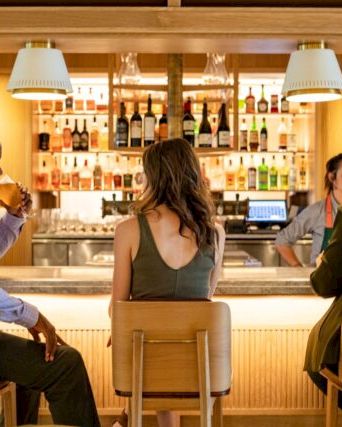 A bar scene with three people seated, engaging in conversation with a bartender in a warmly lit space, featuring shelves of various drinks.