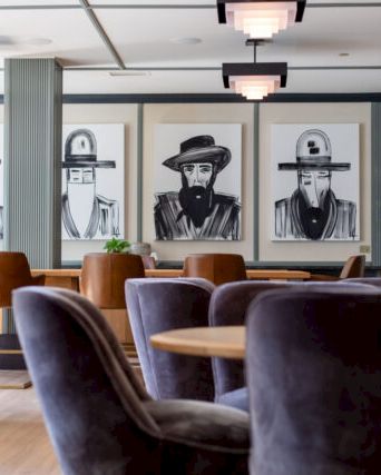 The image shows a modern, stylish lounge with plush chairs, wall art of figures, bookshelves, and soft lighting.