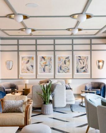 The image shows a modern, stylish lounge with comfortable seating, artwork on the walls, and natural light streaming in from large windows.