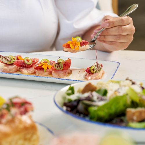 A person in white top eating a plated bite-sized appetizer, with a salad bowl and a pink drink in the foreground, in a cozy dining setting.