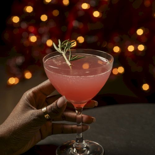 A pink cocktail with a rosemary garnish, held in a hand on a dark table, warm bokeh lights in the background.