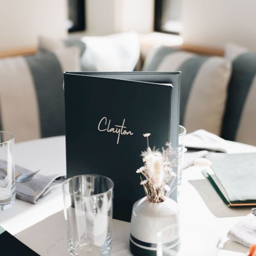 A stylish dining setup with a dark menu card, glassware, napkins, and a small centerpiece on a bright table.