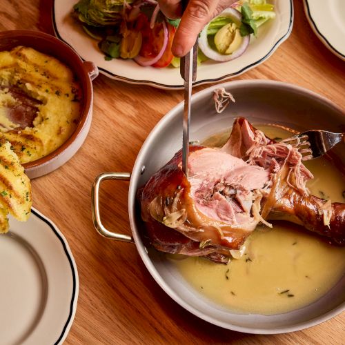 A table with skewers and a bone-in ham or meat dish in a sauce, being cut by forks, with a side dish and plates nearby.
