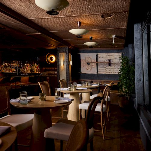 A cozy, dimly lit restaurant with round tables set for dining, warm wood tones, and soft overhead lighting. The scene feels intimate and inviting.