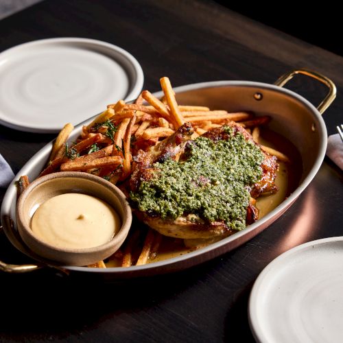 A pan of roasted chicken topped with herbs, served with crispy fries and a side of dipping sauce on a table with plates and utensils.