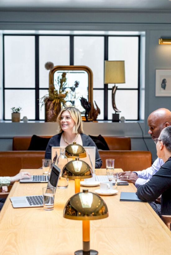 A group of five people sit around a table in a well-decorated meeting room, each with laptops, engaged in discussion.