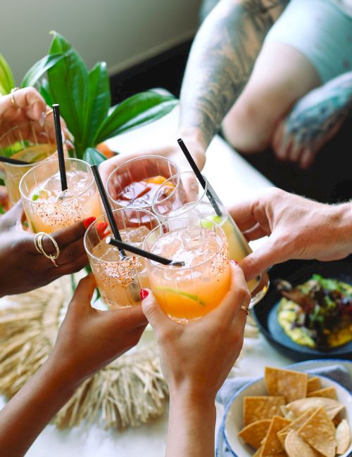 Four people toasting with colorful cocktails, surrounded by dishes with chips and avocado. Tattooed arms are visible.