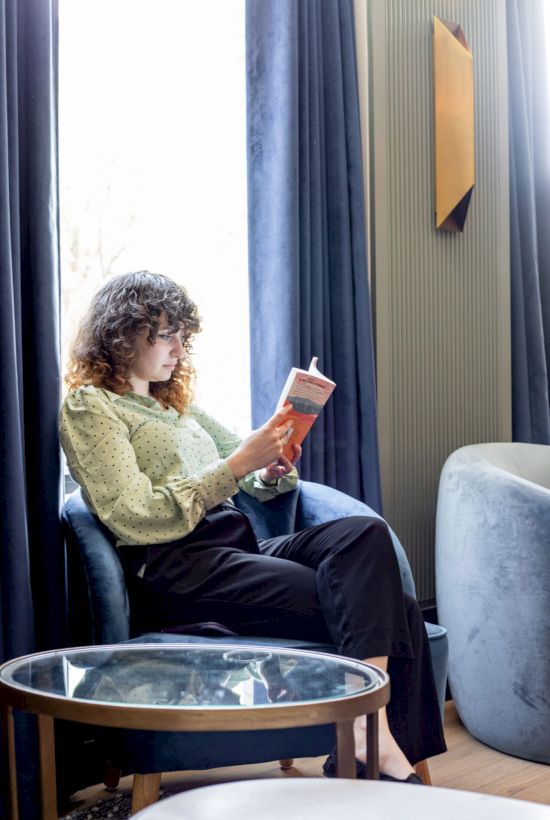 A person with curly hair is sitting on a blue chair by a window, reading a book in a cozy room with blue curtains and a small round table.