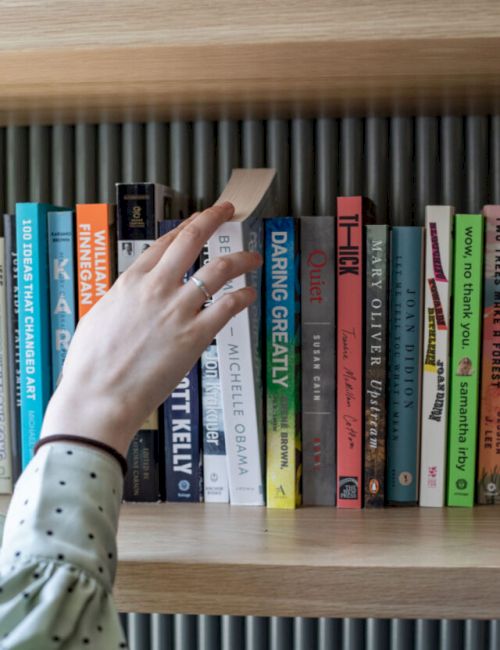 A hand reaching for a book on a shelf, surrounded by various titles, including "Daring Greatly" and "Becoming" by Michelle Obama.