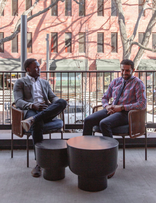 Two people are seated outdoors at a table with three stools, having a conversation against an urban backdrop featuring windows and trees.