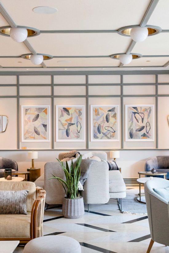 A modern lounge features plush seating, artwork on walls, and large windows, creating a cozy yet elegant ambiance with a touch of greenery.
