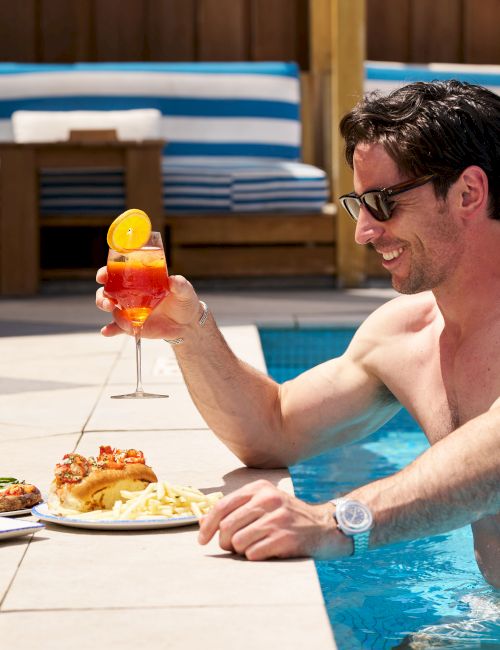 A man in a pool holds a drink while enjoying a plateful of food beside him, with lounge chairs in the background.