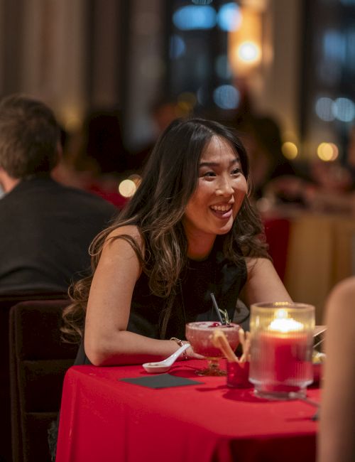 Two people chat warmly at a candlelit restaurant; a smiling woman with long dark hair sits at a red table, lit by candles, others in the background.