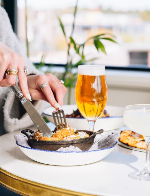 A person cuts into a dish at a sunny table&mdash;beer, chips, and a lime-topped drink nearby, with additional plates in view.