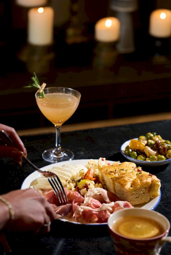 A person&rsquo;s meal with bread, cured meat, salad, and a cocktail&mdash;bar setup with candles in the background. End of sentence.