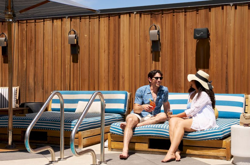 Two people sit on a striped lounge by a pool, chatting, with a wooden privacy wall and clear blue sky behind them.
